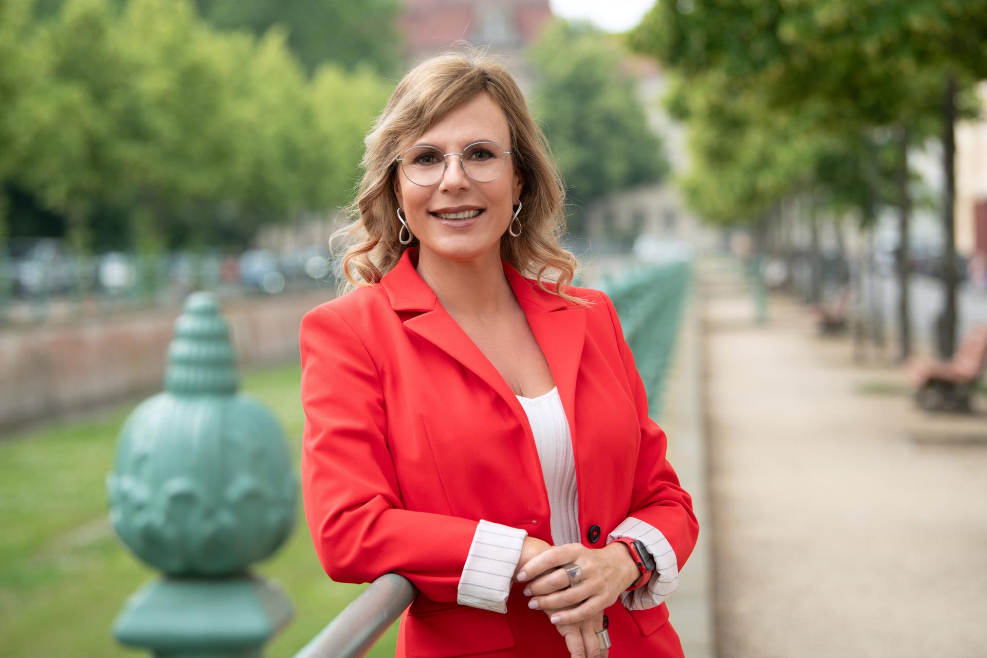 Anika Tannebaum – Europe’s No. 1 service expert, keynote speaker, and leadership coach, smiling in a red blazer on a bridge