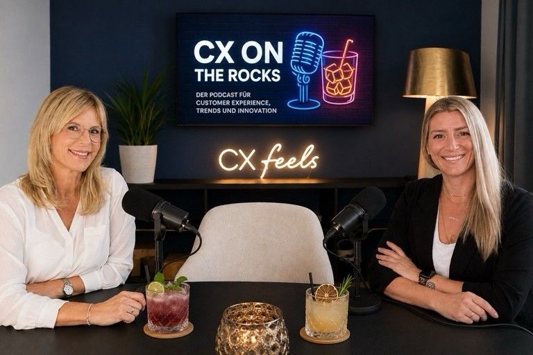Anika Tannebaum und Annika Schmidt sitzen lächelnd an einem Tisch mit Cocktails und Mikrofonen, im Hintergrund ein Leuchtschild "CX on the Rocks – Der Podcast für Customer Experience, Trends und Innovation" und der Schriftzug "CX feels"