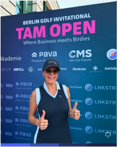 Anika Tannebaum in dunkelblauem Golf-Outfit mit Cap zeigt lachend beide Daumen hoch vor der Fotowand des "Berlin Golf Invitational – TAM OPEN"